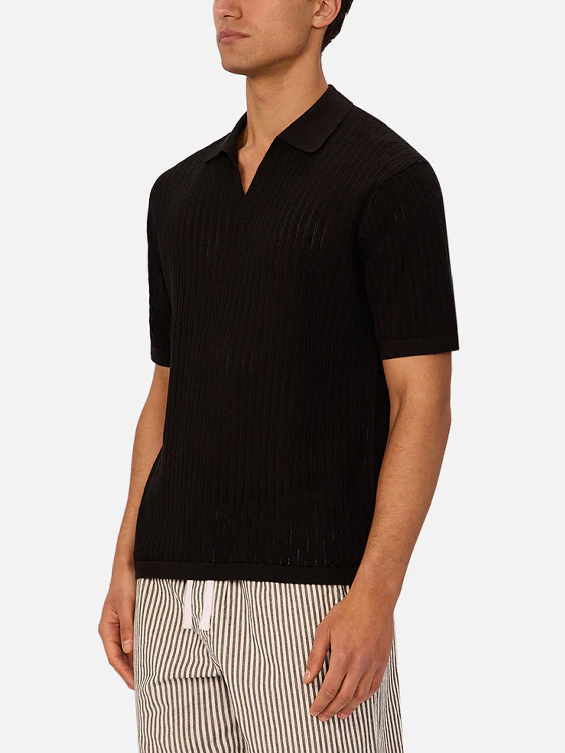 A man wears the Industrie Poolside Polo, a black short-sleeve knit shirt, paired with light shorts with vertical black stripes, standing against a plain light background.