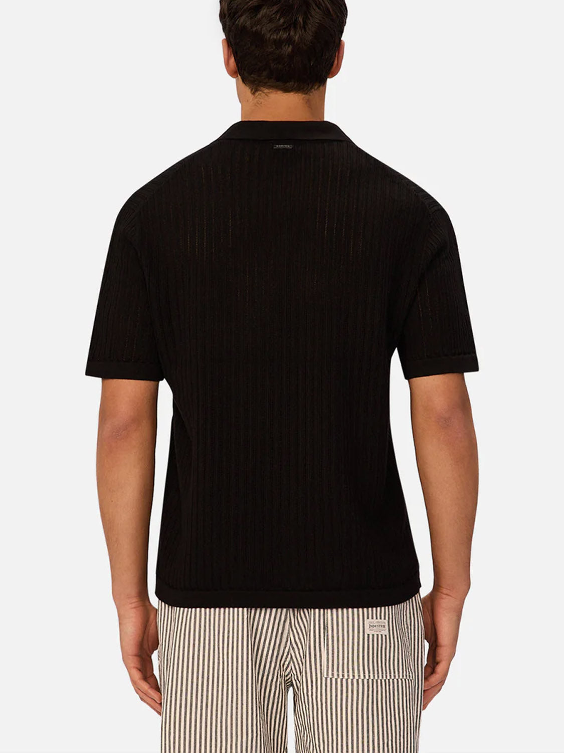 A man is seen from behind wearing the Industrie Poolside Polo, a short-sleeved, black ribbed knit shirt, paired with light striped shorts with a back pocket. The scene is set against a plain white background.