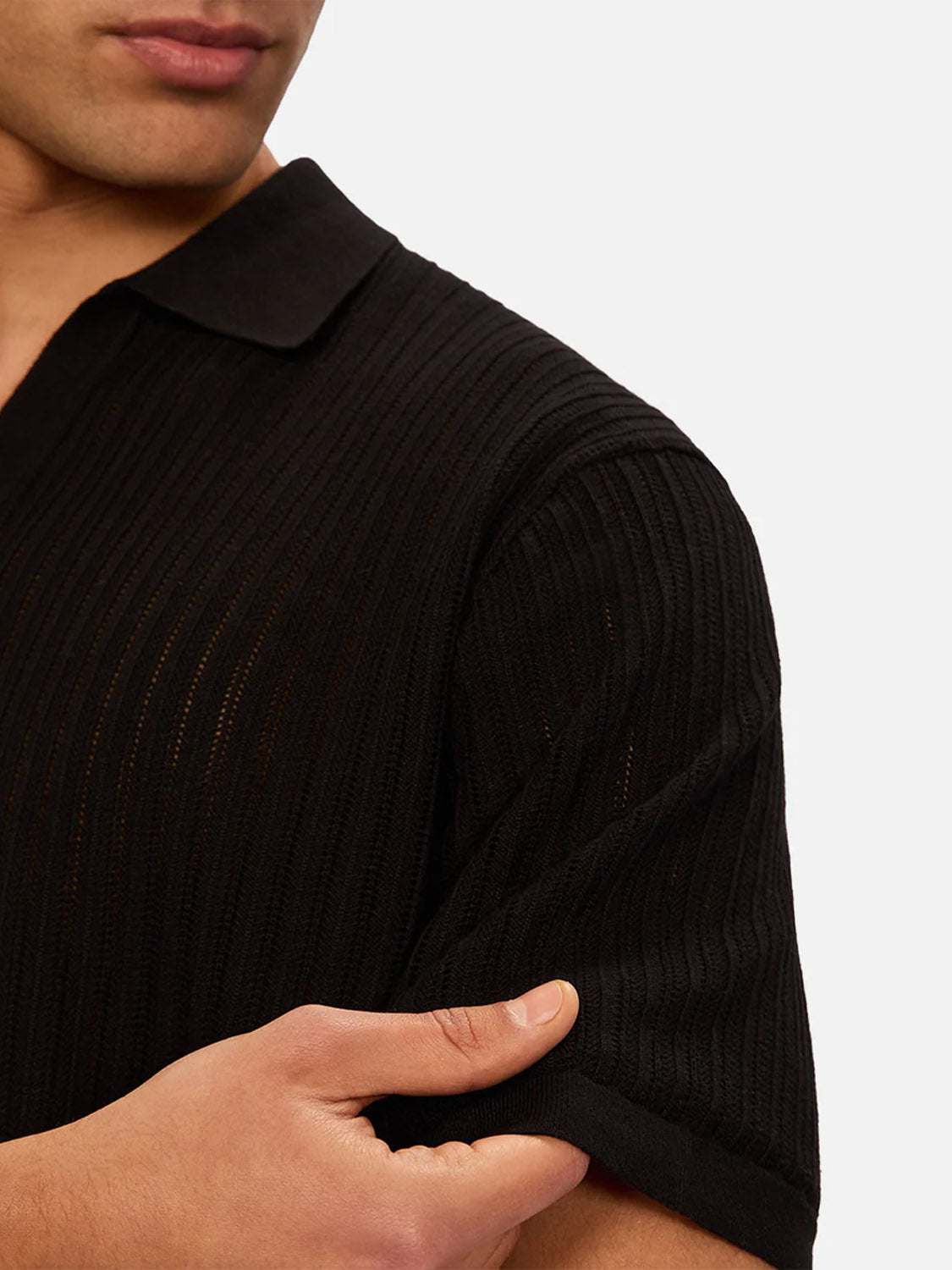 Wearing the Industrie Poolside Polo in black, a person rolls up the short sleeve with one hand; only their lower face, neck, and upper torso appear against a plain white background.