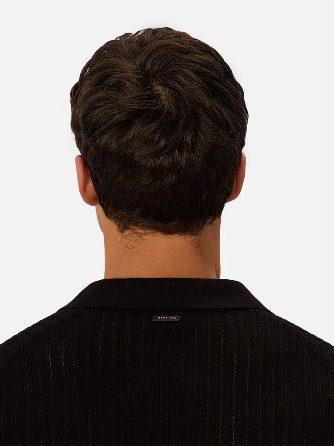 A man with short brown hair is seen from behind wearing the Industrie Poolside Polo, a black ribbed cotton shirt with a small rectangular label near the collar. The background is plain and light-colored.