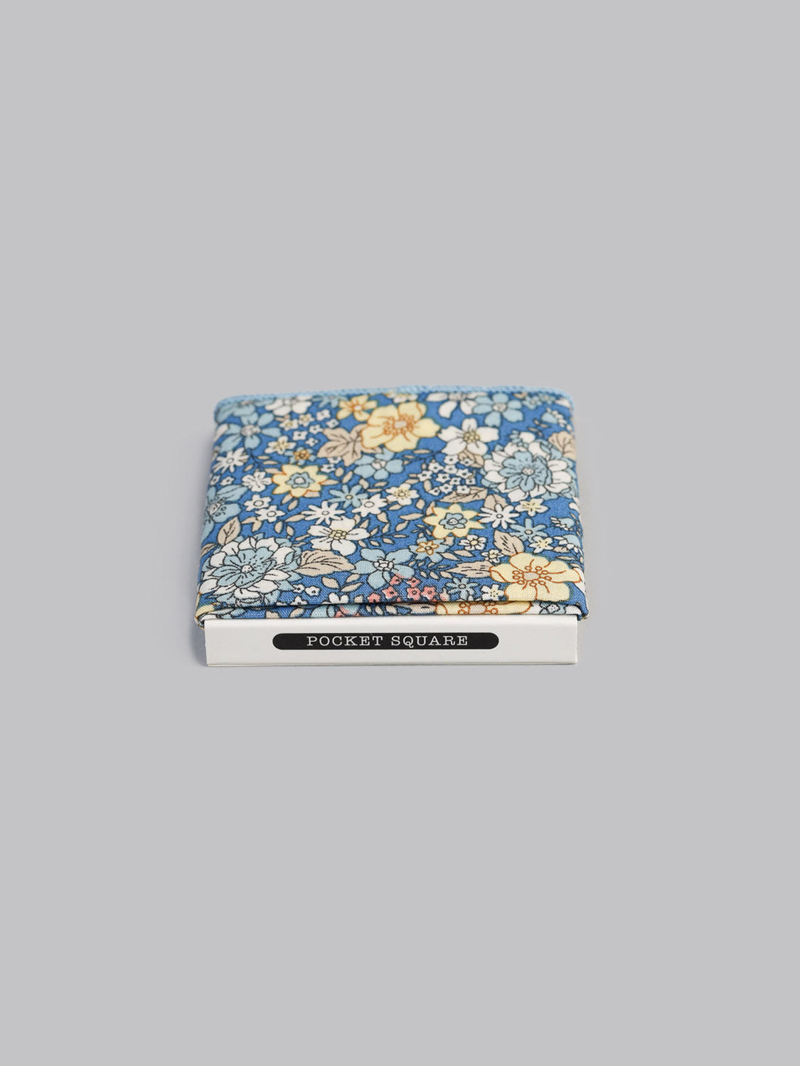 The Poplin Pocket Square by ID Menswear is a blue cotton accessory with a yellow and white floral pattern, handmade in Brooklyn, neatly folded in a white POCKET SQUARE box on a light gray background.