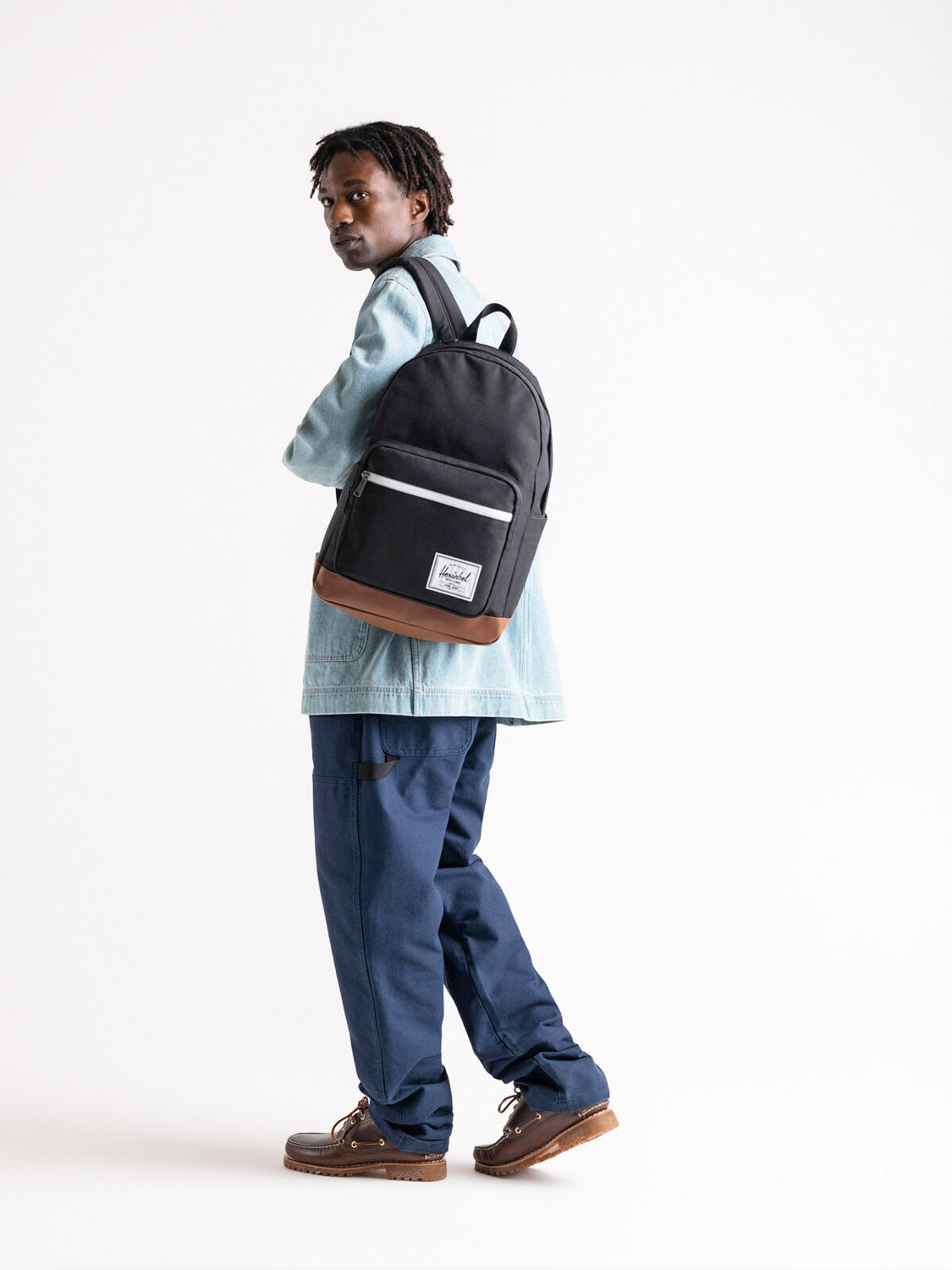 A person stands facing slightly away, looking over their shoulder, wearing a black Herschel Pop Quiz Backpack with brown trim and a white label, crafted from EcoSystem fabric, against a plain white background.