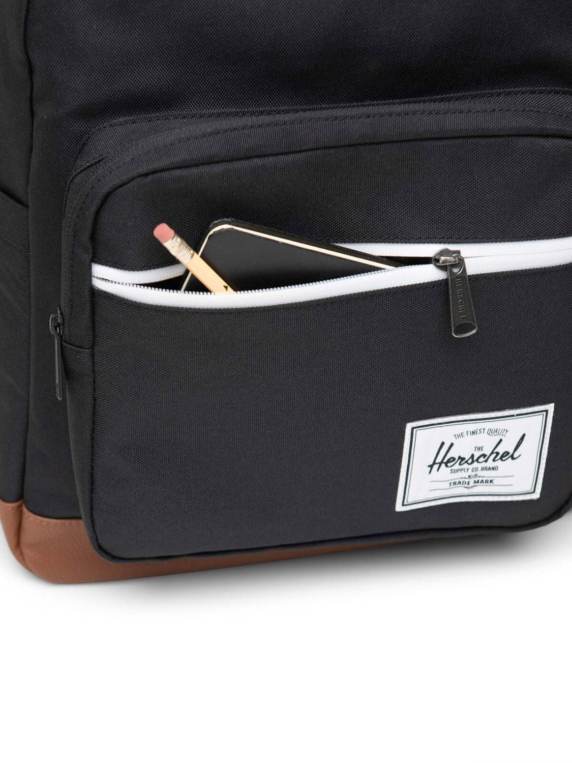 A close-up of the Herschel Pop Quiz Backpack in black with a brown bottom made from EcoSystem fabric. The front pocket is partially unzipped to reveal a pencil and smartphone, with the Herschel label visible on the pocket.