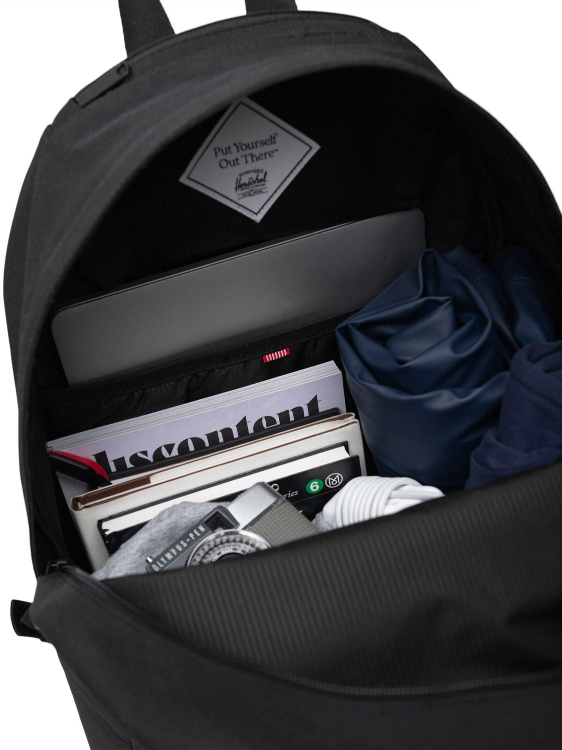 An open black Herschel Pop Quiz Backpack made from EcoSystem fabric reveals a laptop, magazines, a notebook, folded clothes, and a watch inside. A label reads "Put Yourself Out There.