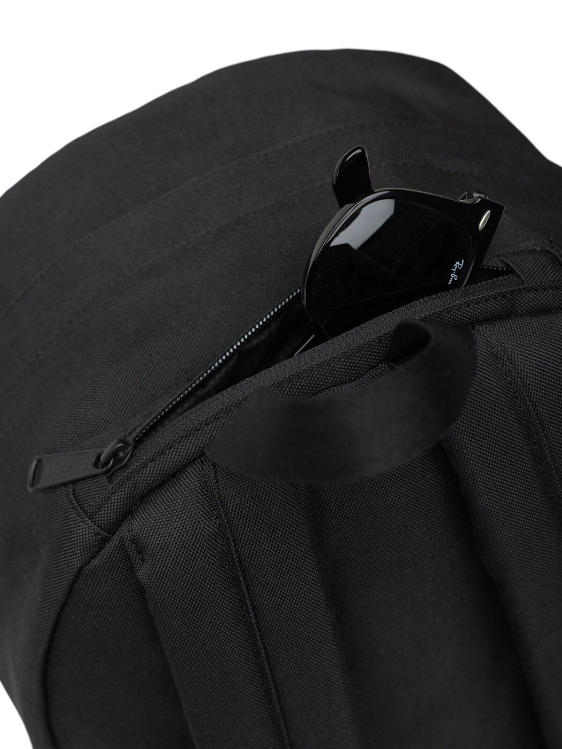A pair of black sunglasses partially tucked into the zippered pocket of a black Herschel Pop Quiz Backpack made from EcoSystem fabric, shown against a white background.