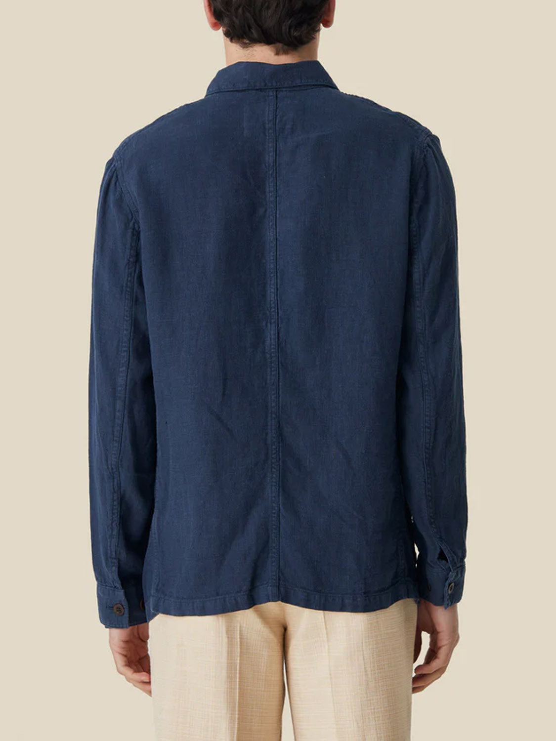 Rear view of a person wearing a Portuguese Flannel Labura Linen Jacket in dark blue and light beige pants, standing against a beige background. The jacket has a simple, casual design with a collared neckline, reflecting the timeless elegance of Portuguese heritage.