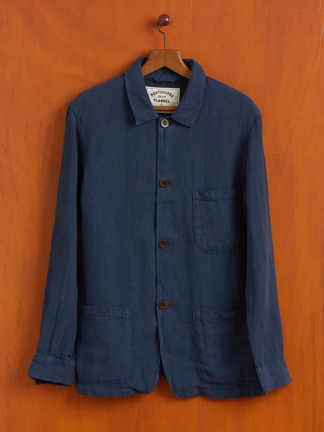 A navy blue Portuguese Flannel Labura Linen Jacket with a collar hangs on a wooden wall, reflecting Portuguese heritage. It includes two large front pockets and one chest pocket, dark buttons, and a label that reads “Portuguese Flannel,” ensuring craftsmanship from quality mills.