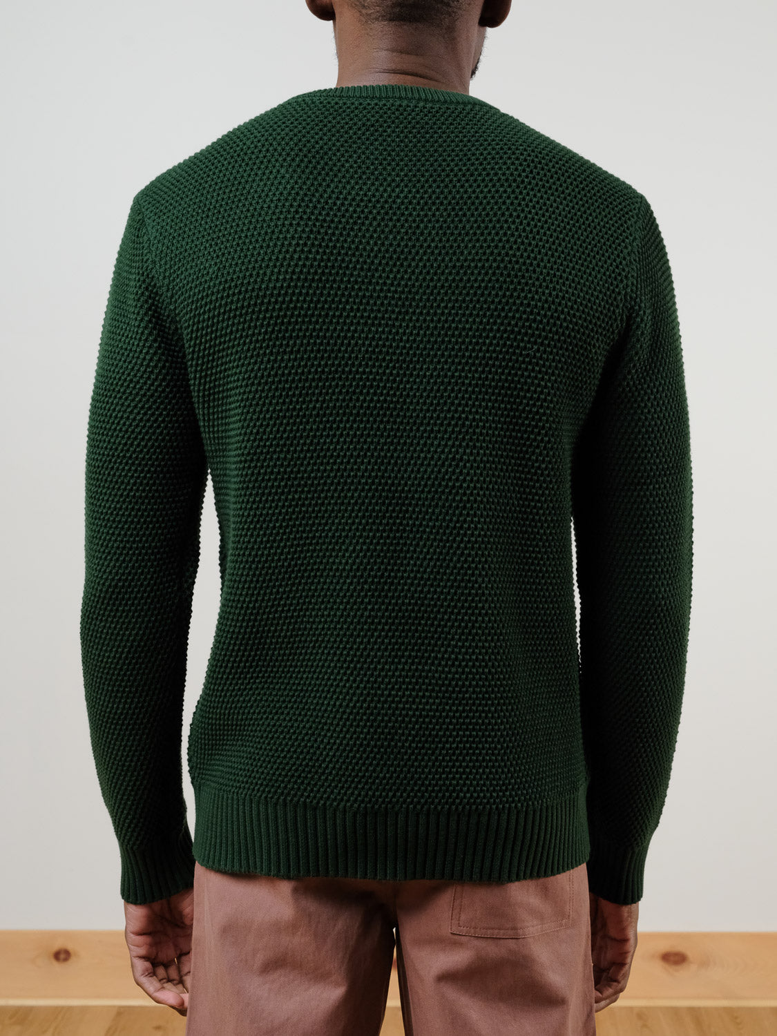 A person stands facing away from the camera, wearing the Quebec Chunky Knit Sweater by ID Menswear in textured dark green and light brown pants, against a plain light-colored wall and light wooden floor.