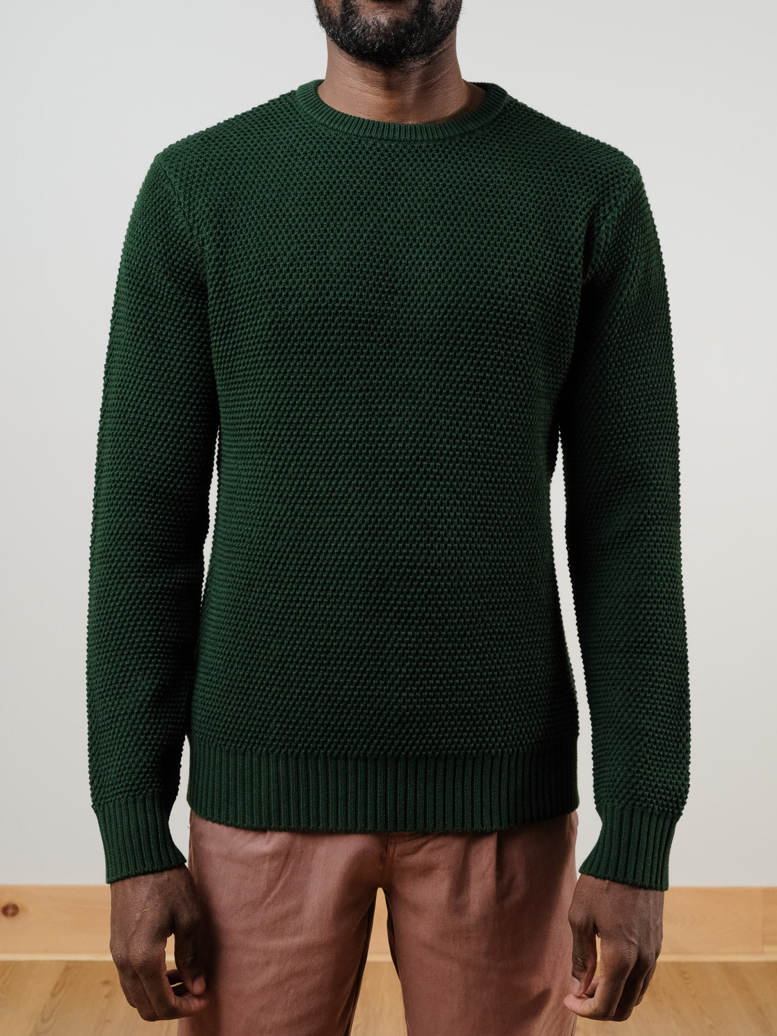 A person stands indoors against a plain light background wearing the Quebec Chunky Knit Sweater by ID Menswear in dark green, paired with light brown pants. The individual&#39;s head is partially out of frame.