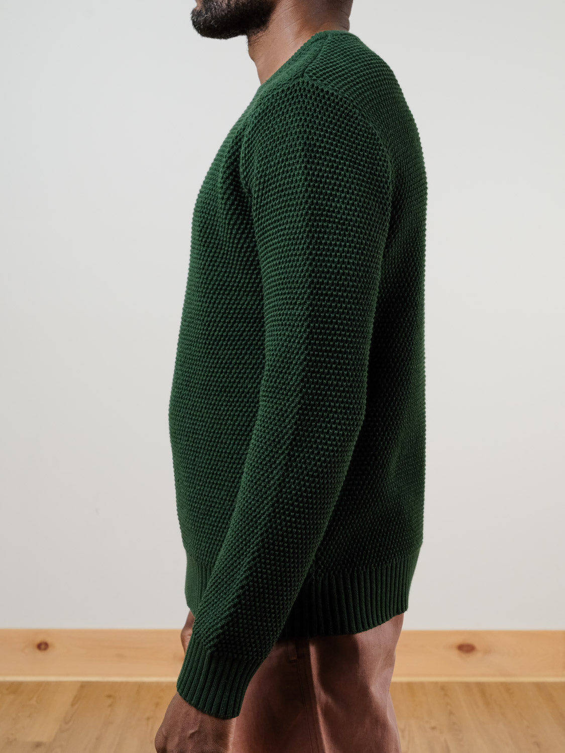 A man stands in profile against a plain background with a wooden floor, wearing the ID Menswear Quebec Chunky Knit Sweater in dark green moss and light brown pants.