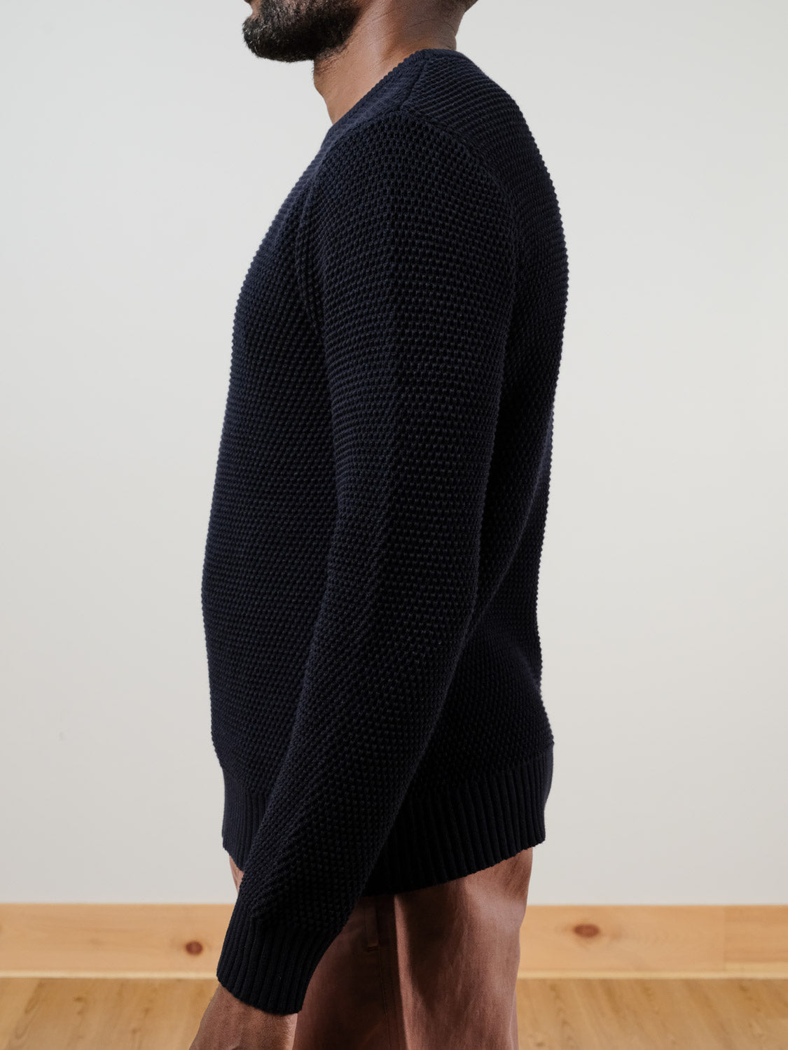A man stands in profile wearing the Quebec Chunky Knit Sweater by ID Menswear in navy, paired with light brown pants. He’s set against a plain backdrop with a light wall and visible wooden floor trim.