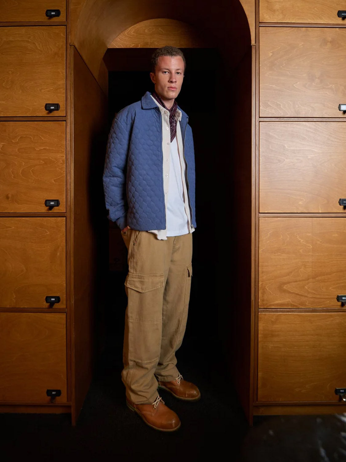 A man stands in a wooden hallway wearing the Kuwalla Quilted Corduroy Shacket—featuring a front zipper—over a white shirt and tan cargo pants, with built-in wood cabinets and an arched doorway in the background.