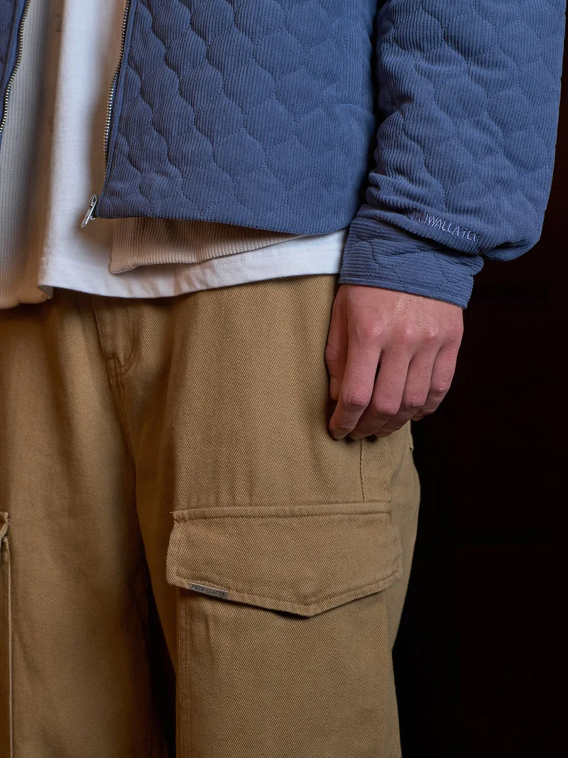 A person wearing the Kuwalla Quilted Corduroy Shacket, featuring a front zipper closure, over a white shirt and tan cargo pants; only their lower torso and right hand are visible against a dark background.