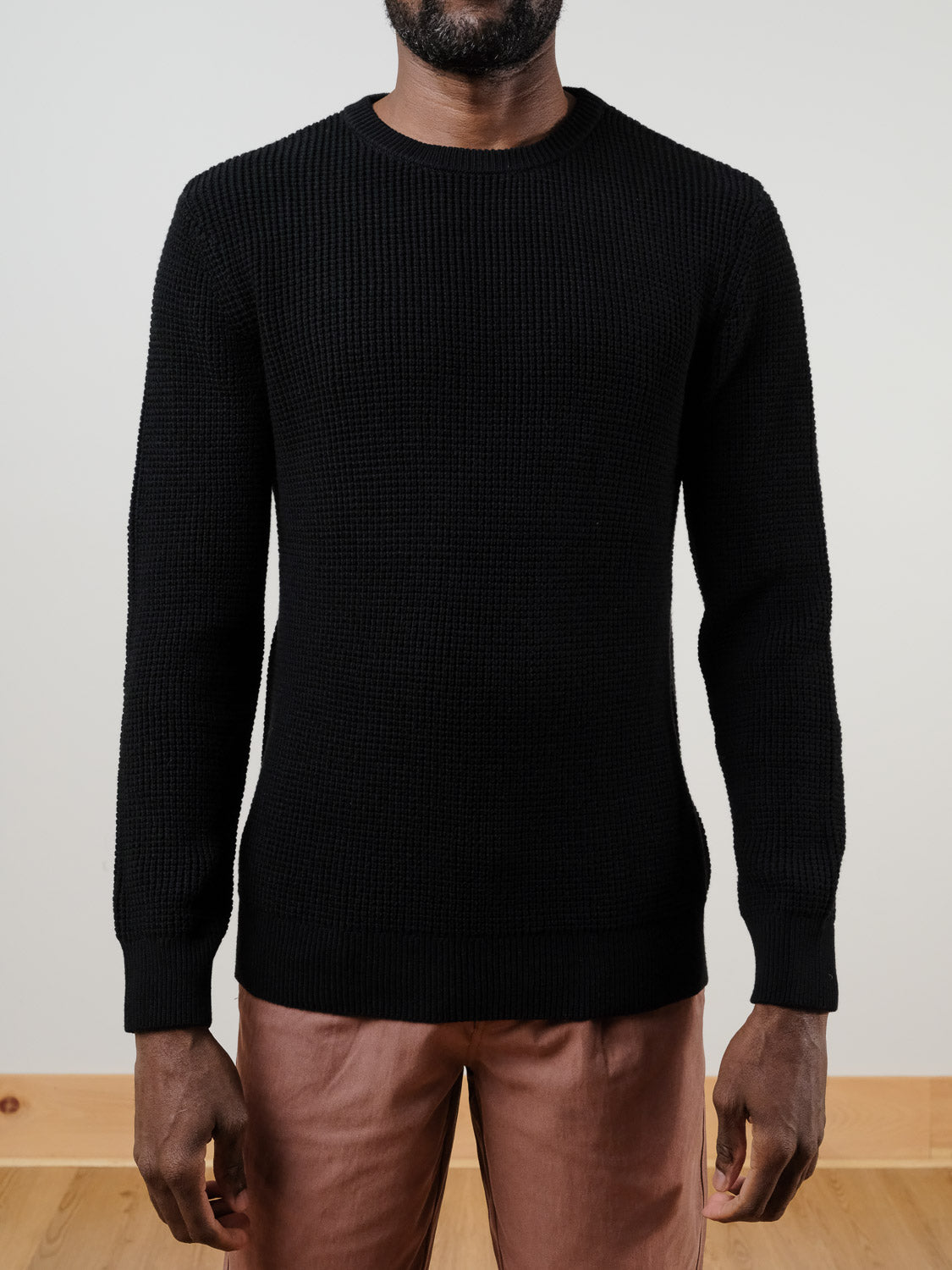 A man wears the Ragnar Waffle Knit Sweater by ID Menswear in textured black 100% cotton, paired with light brown pants. The waffle knit detail is visible in this cropped photo against a plain wall and wooden floor.