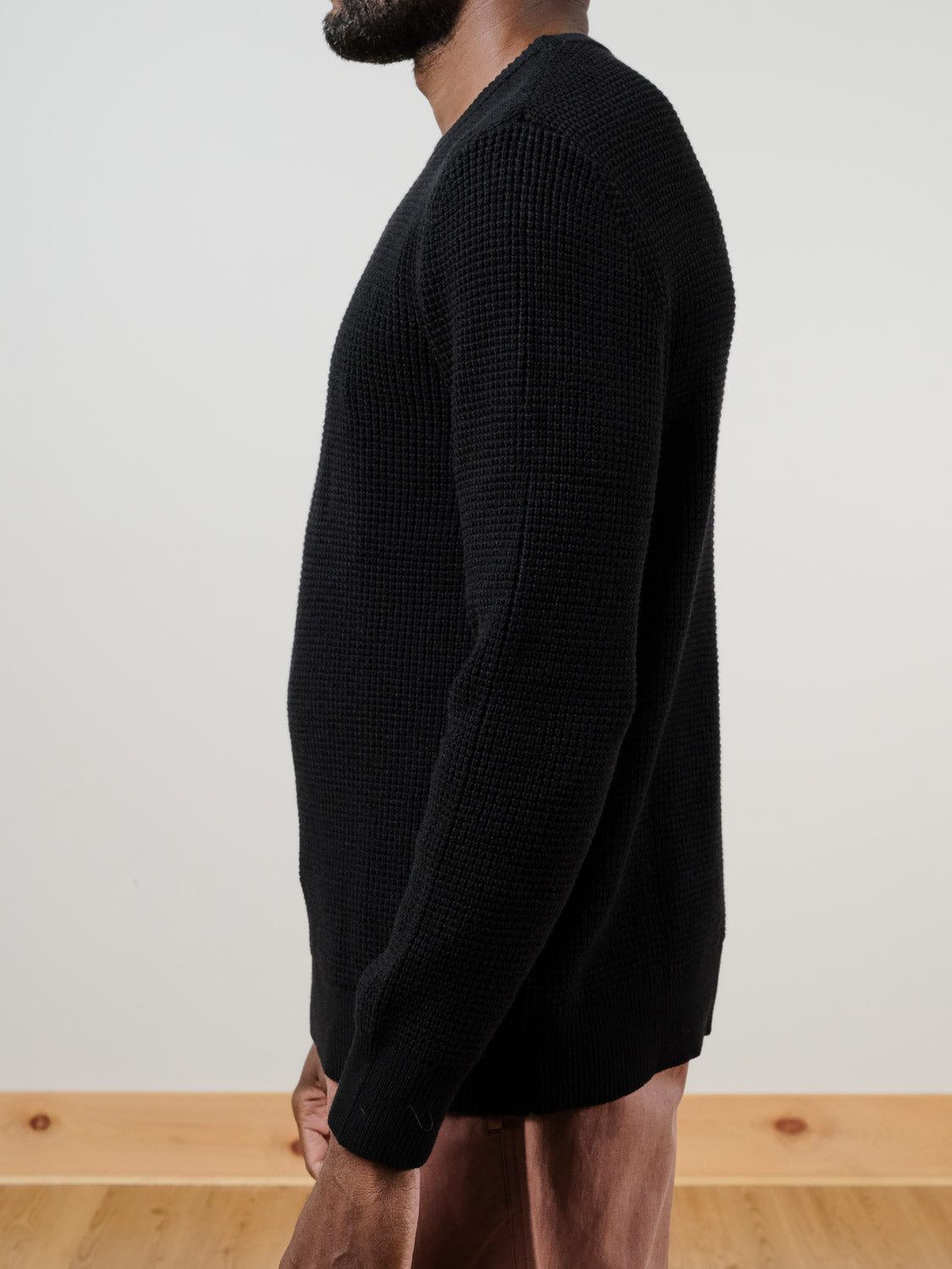 A man stands in profile against a light wall and wooden floor, wearing the Ragnar Waffle Knit Sweater by ID Menswear in black 100% cotton with brown pants. His face is mostly out of frame.