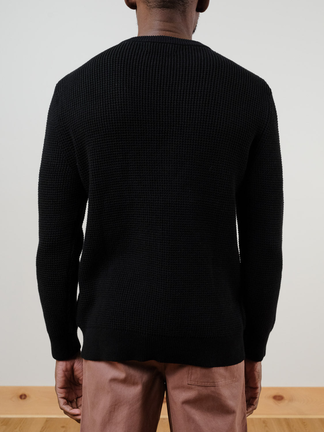 A person faces away from the camera, wearing the black Ragnar Waffle Knit Sweater by ID Menswear and light brown pants, against a plain, minimally decorated background.