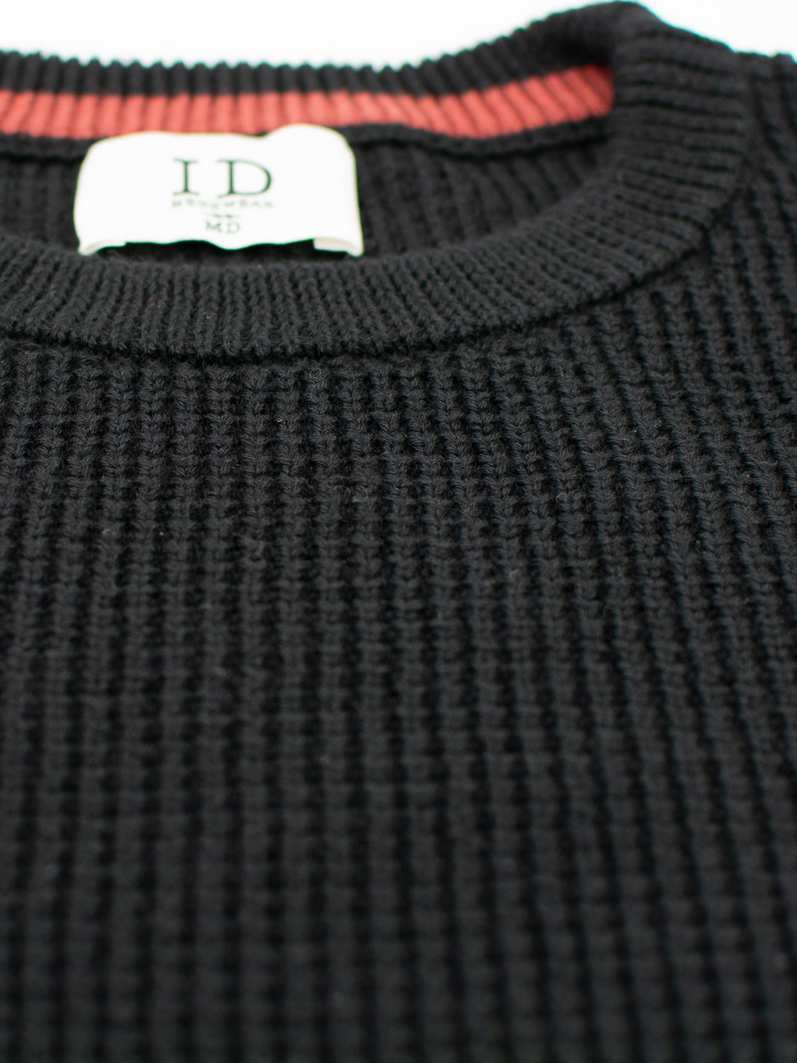This close-up showcases the Ragnar Waffle Knit Sweater from ID Menswear, featuring a black round neck design. The red seam on the inside collar and the white label enhance its style. Made from 100% cotton, it highlights the waffle knit texture beautifully.