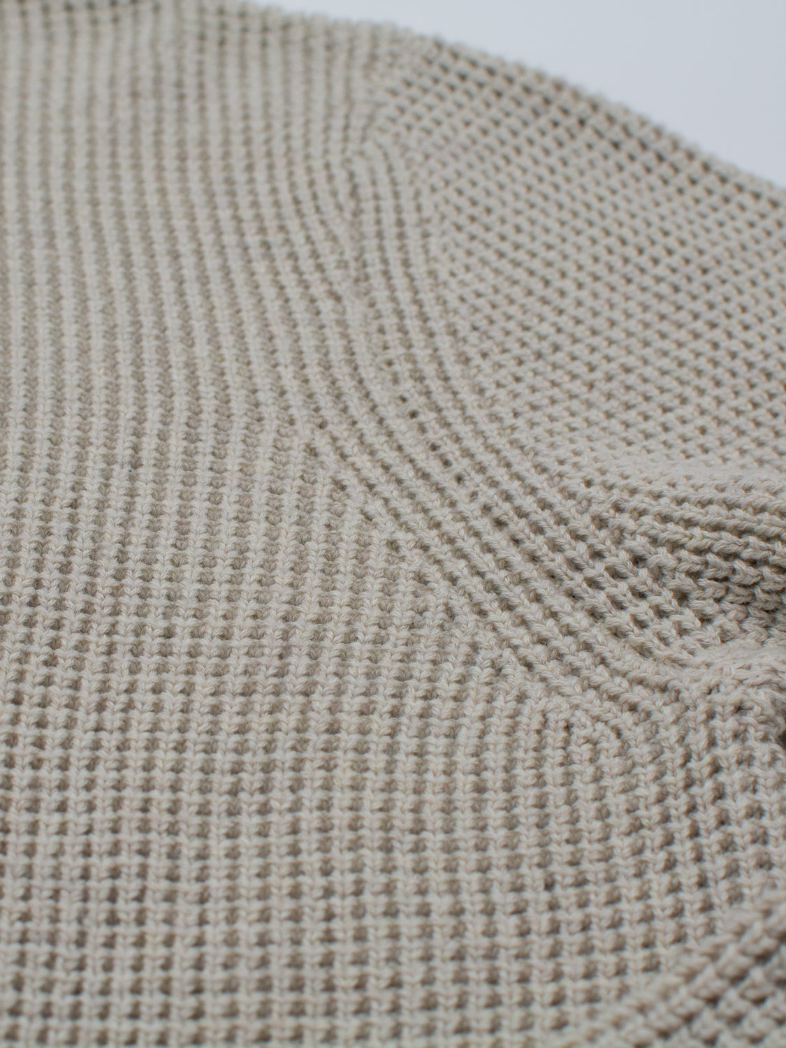 A close-up of a light beige fabric reveals a chunky, ribbed waffle knit texture reminiscent of the Ragnar Waffle Knit Sweater by ID Menswear. The fibers form a cozy, soft pattern, suggesting flexibility and are crafted from 100% cotton.