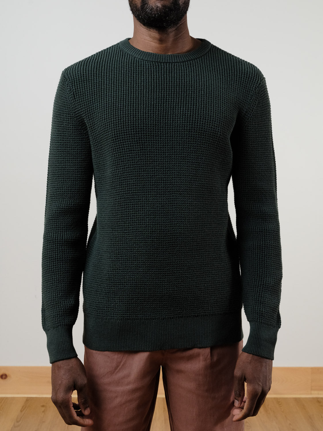 A person wears the ID Menswear Ragnar Waffle Knit Sweater in textured dark green 100% cotton and brown pants, standing against a plain light wall and wooden floor with arms relaxed at their sides. Their head is partially out of frame.