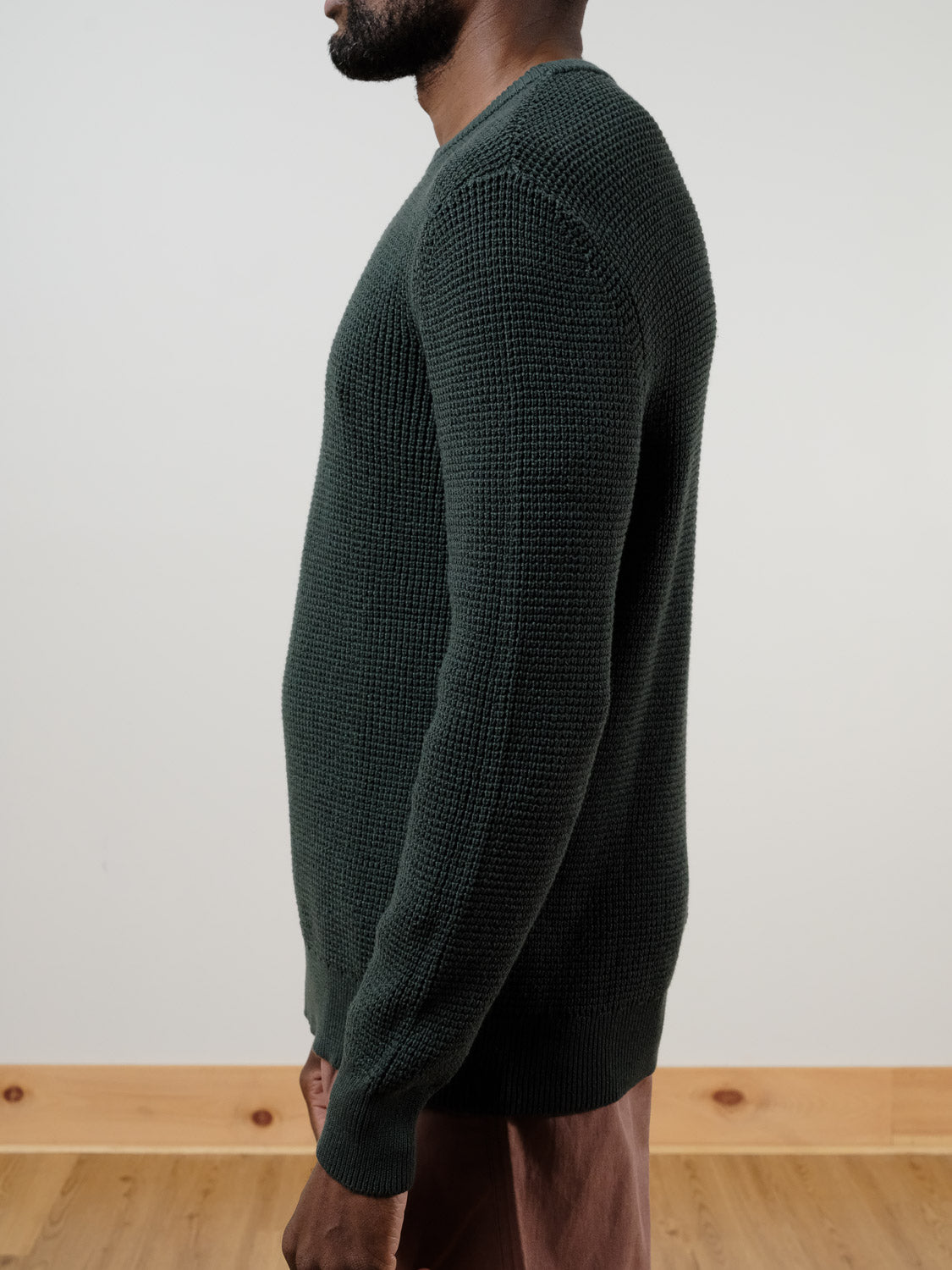 A person stands in profile against a light wall and wooden floor, wearing the ID Menswear Ragnar Waffle Knit Sweater in textured dark green cotton, paired with brown pants.