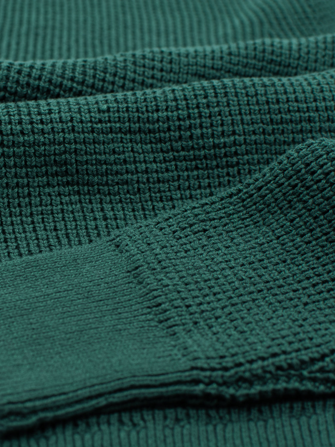 Close-up of a dark green waffle-textured fabric, similar to ID Menswears Ragnar Waffle Knit Sweater. The folded material highlights its deep shadows and soft, knitted 100% cotton structure.