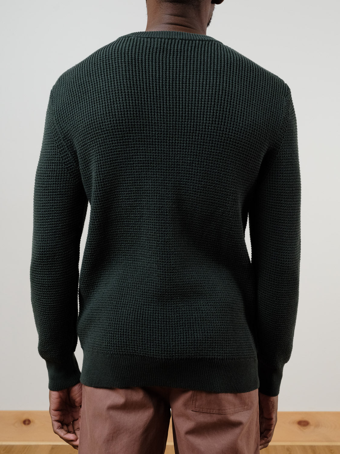 A person stands facing away, wearing the ID Menswear Ragnar Waffle Knit Sweater in dark green 100% cotton and light brown pants, set against a plain indoor background with a wooden floor.
