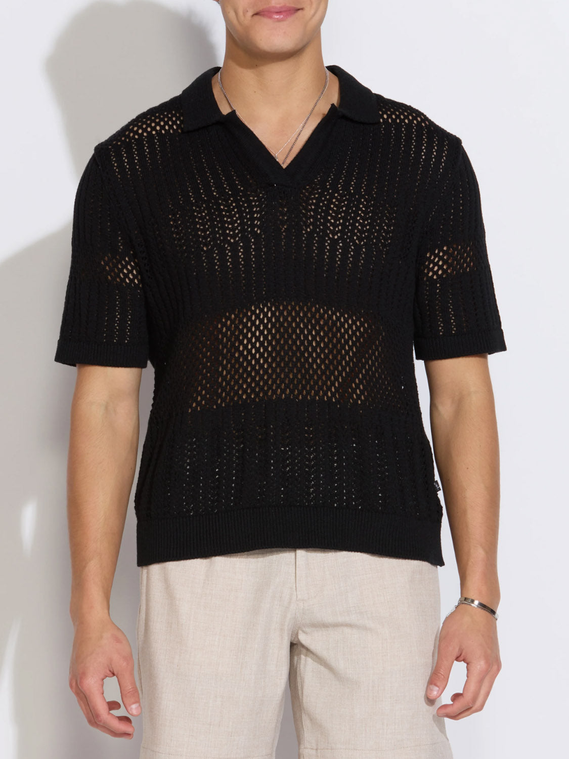 A man wears the Civil Society Rapa Boxy Fit Johnny Collar Crochet Sweater Polo in black with light beige pants, standing against a white background—a stylish pick for men's summer knitwear.