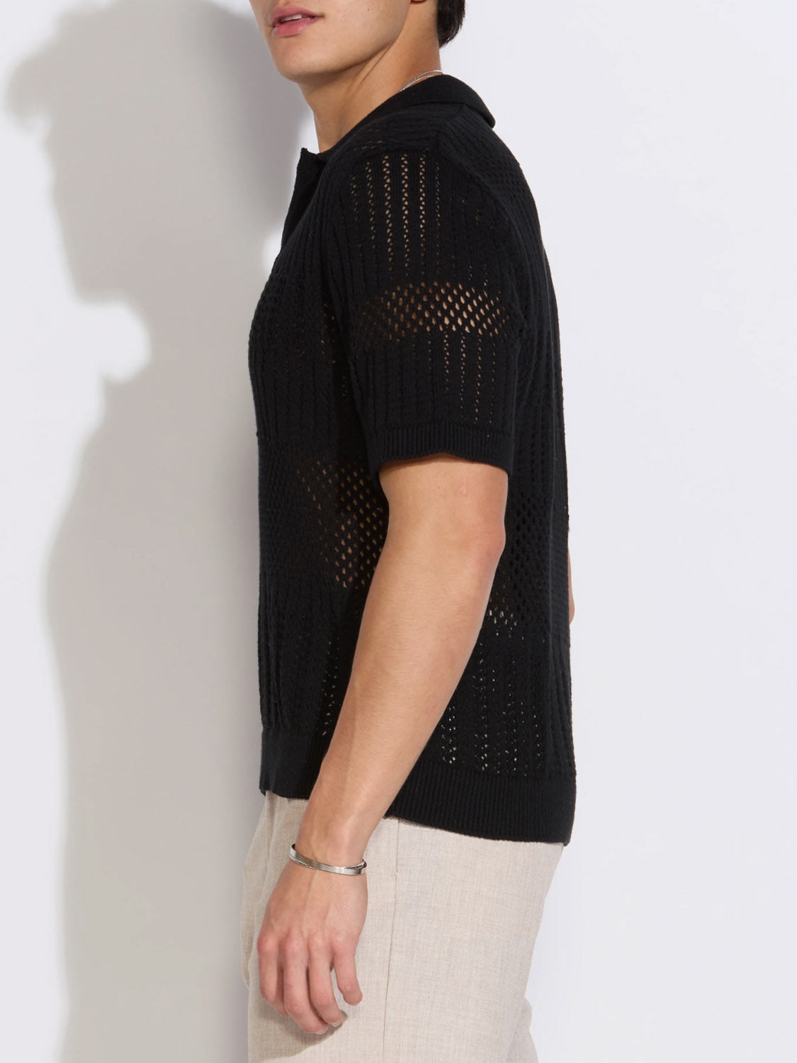 A person in a black Rapa Boxy Fit Johnny Collar Crochet Sweater Polo by Civil Society and light pants stands sideways against a white background, partly out of frame, highlighting stylish men’s summer knitwear.
