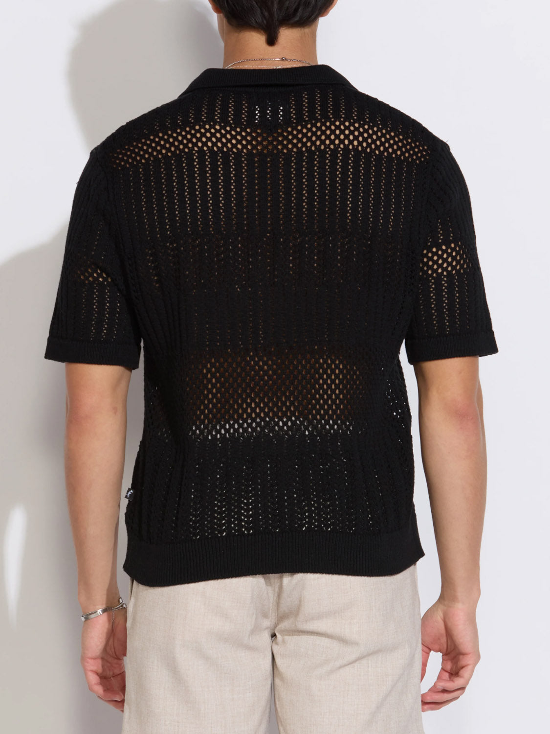 A person is shown from the back wearing Civil Society’s Rapa Johnny Collar Crochet Sweater Polo in black—a 100% cotton, boxy-fit, short-sleeve, open-knit shirt—paired with light beige pants. The patterned see-through polo reveals skin underneath against a plain white background.
