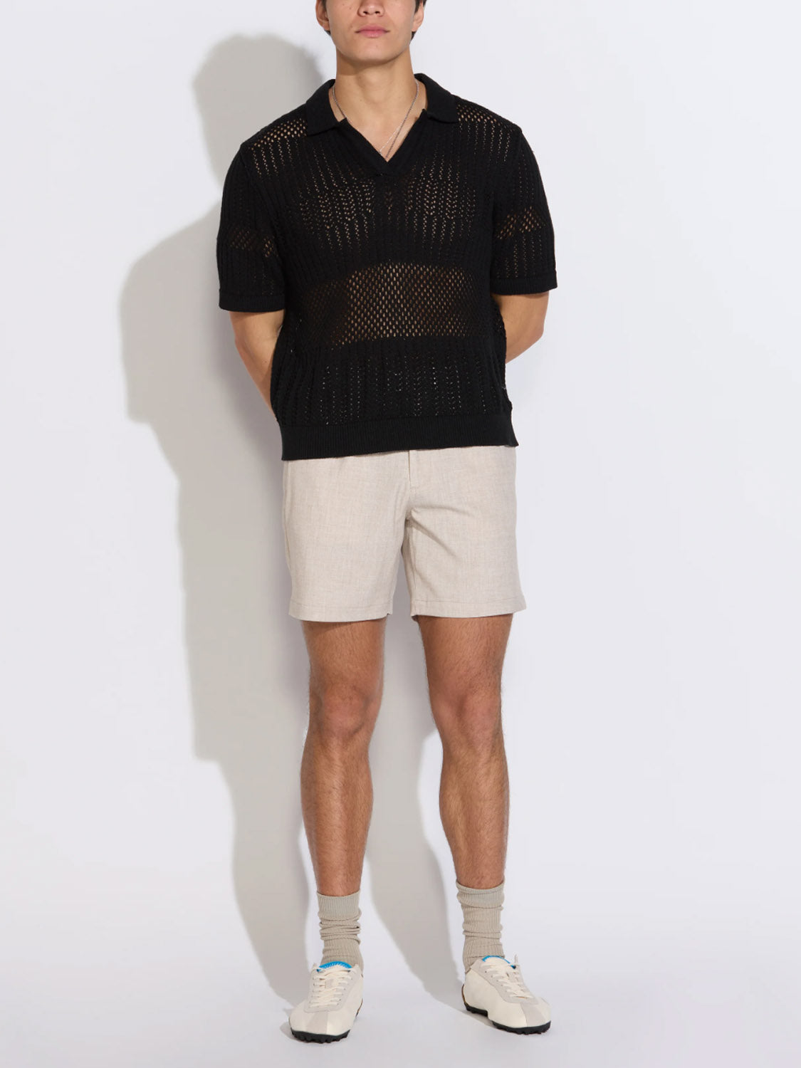 A person wears the Civil Society Rapa Johnny Collar Crochet Sweater Polo in black, beige shorts, light gray socks, and white sneakers, standing against a white background with hands behind their back.