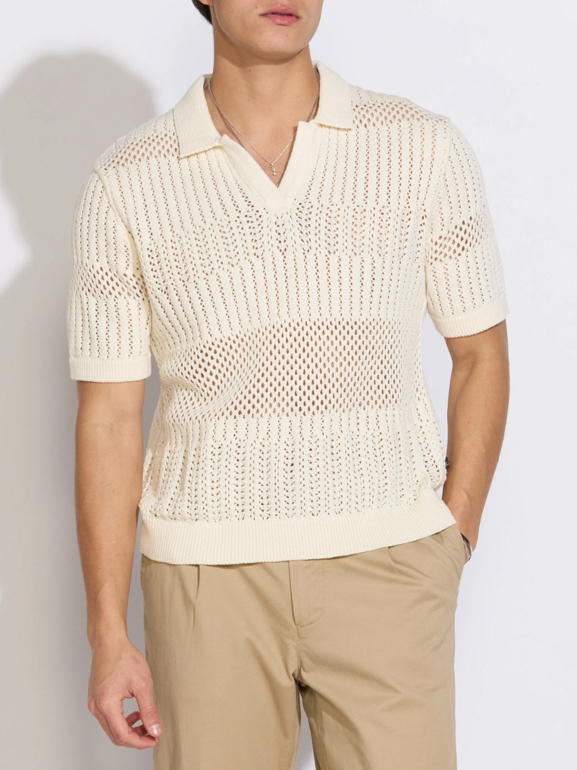 A person wears the Rapa Johnny Collar Crochet Sweater Polo by Civil Society—a cream short-sleeve, boxy-fit sweater with a collar and V-neck—paired with light beige pants, standing against a white background with one hand in their pocket.