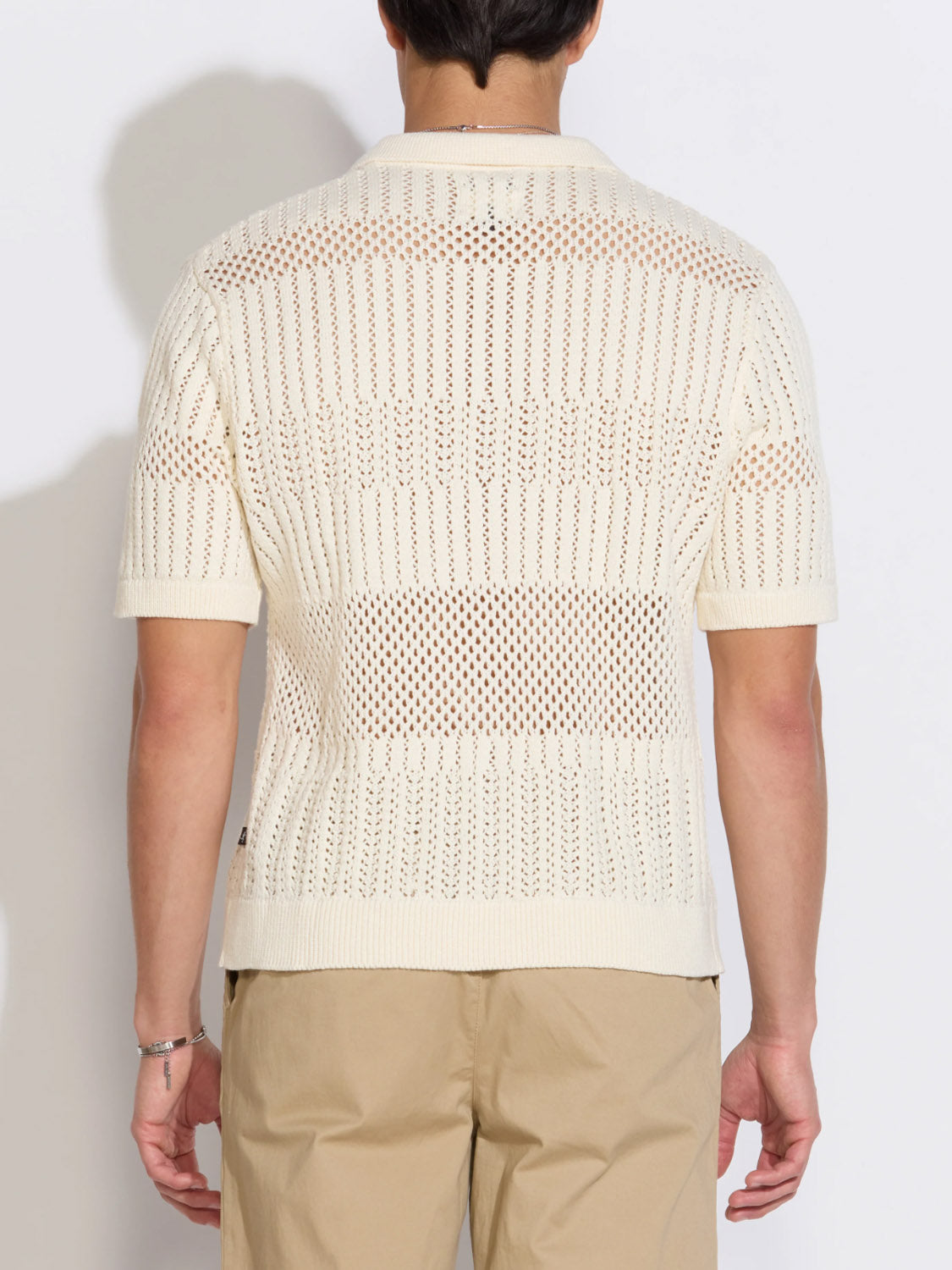 A person is seen from the back wearing Civil Society’s Rapa Johnny Collar Crochet Sweater Polo—a short-sleeve, off-white, 100% cotton top with a boxy fit and mesh-like crochet pattern—paired with light beige pants.