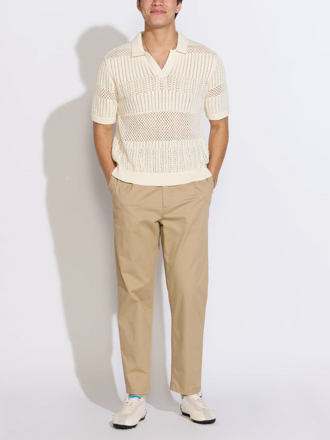 A person stands with hands in pockets, wearing the Civil Society Rapa Johnny Collar Crochet Sweater Polo in cream, featuring short sleeves and a mesh pattern, paired with beige pants and white sneakers against a plain white background.