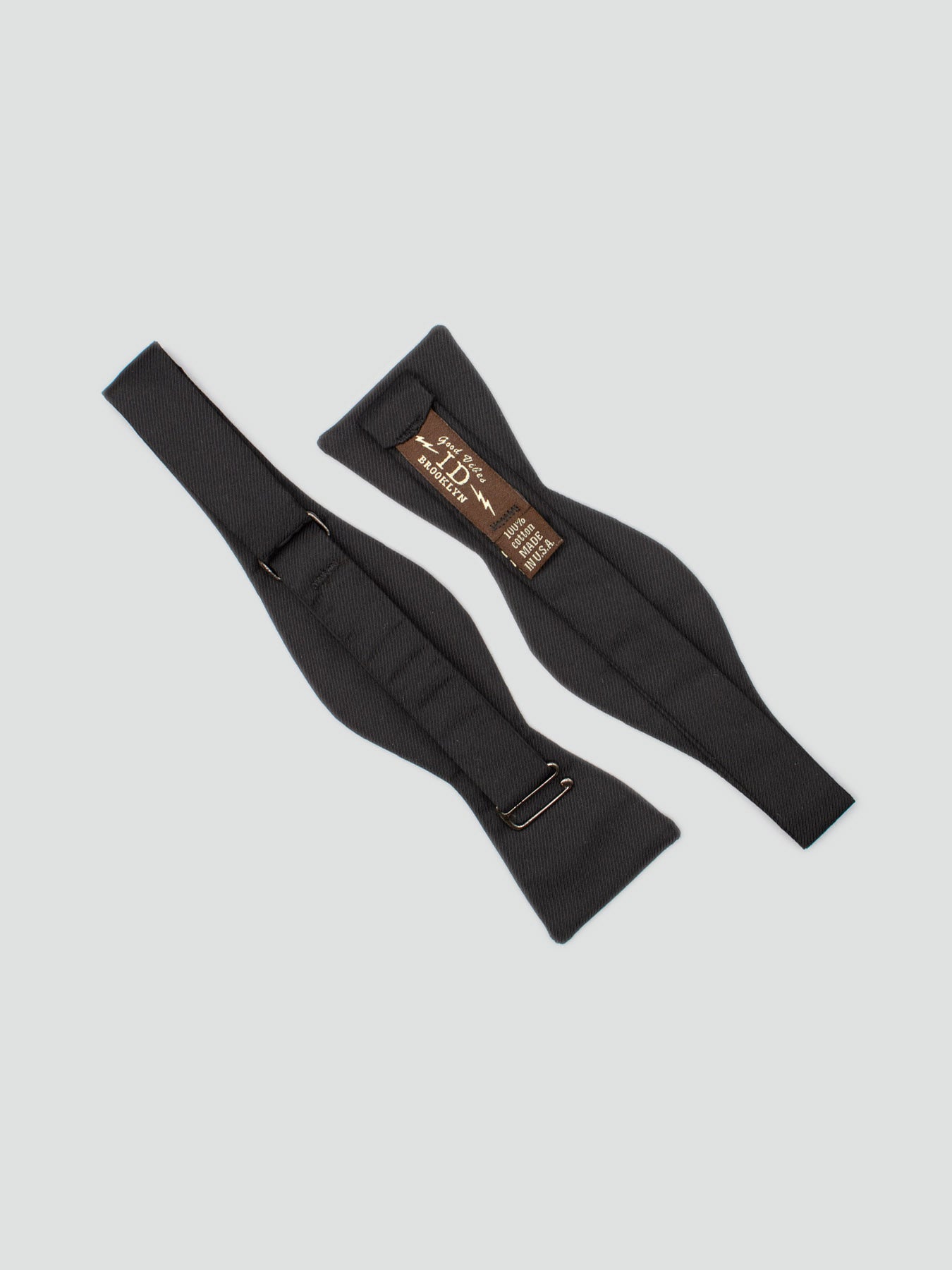 Two Raven Cotton Bow Tie straps from ID Menswear are laid flat on a light grey background—one reveals a brown label with sizing, while the other shows the classic black front, ideal for any formal accessory collection.