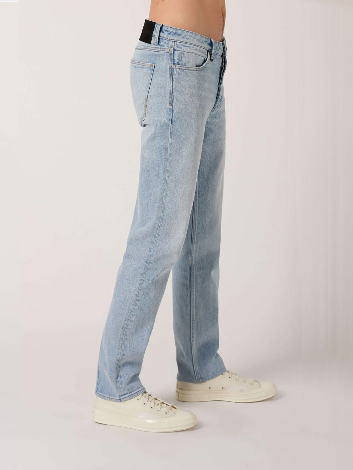 A person wears Neuw Ray Straight Baroque jeans in light blue with off-white sneakers, standing against a plain light background, shown from the waist down in a side profile view.