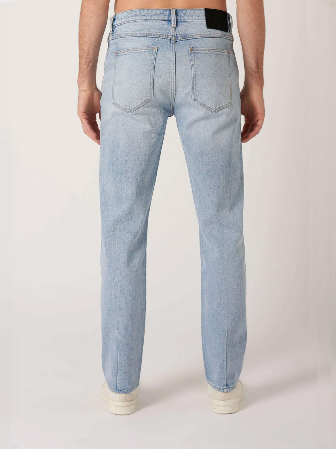 A person is standing with their back to the camera, wearing Neuw Ray Straight Baroque jeans in light blue and white sneakers, set against a plain light background.