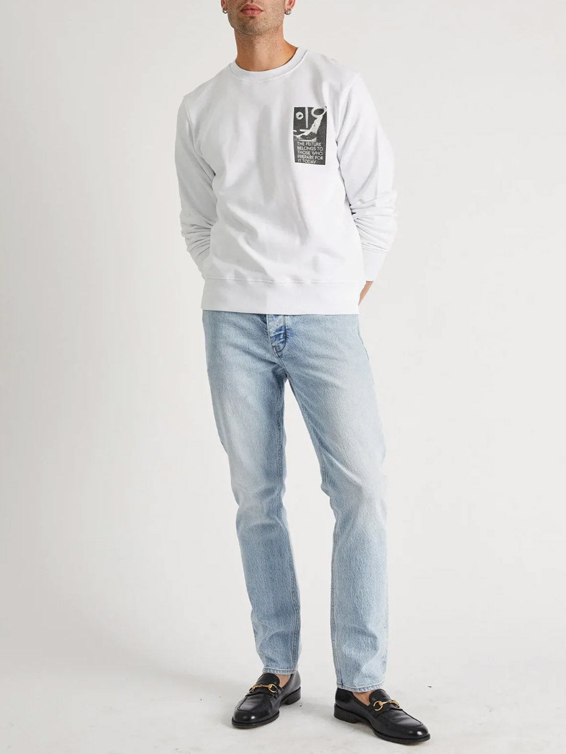 A man stands against a plain white background wearing a white sweatshirt with a black and white graphic patch, Neuw Ray Straight Baroque light blue jeans, and black loafers.