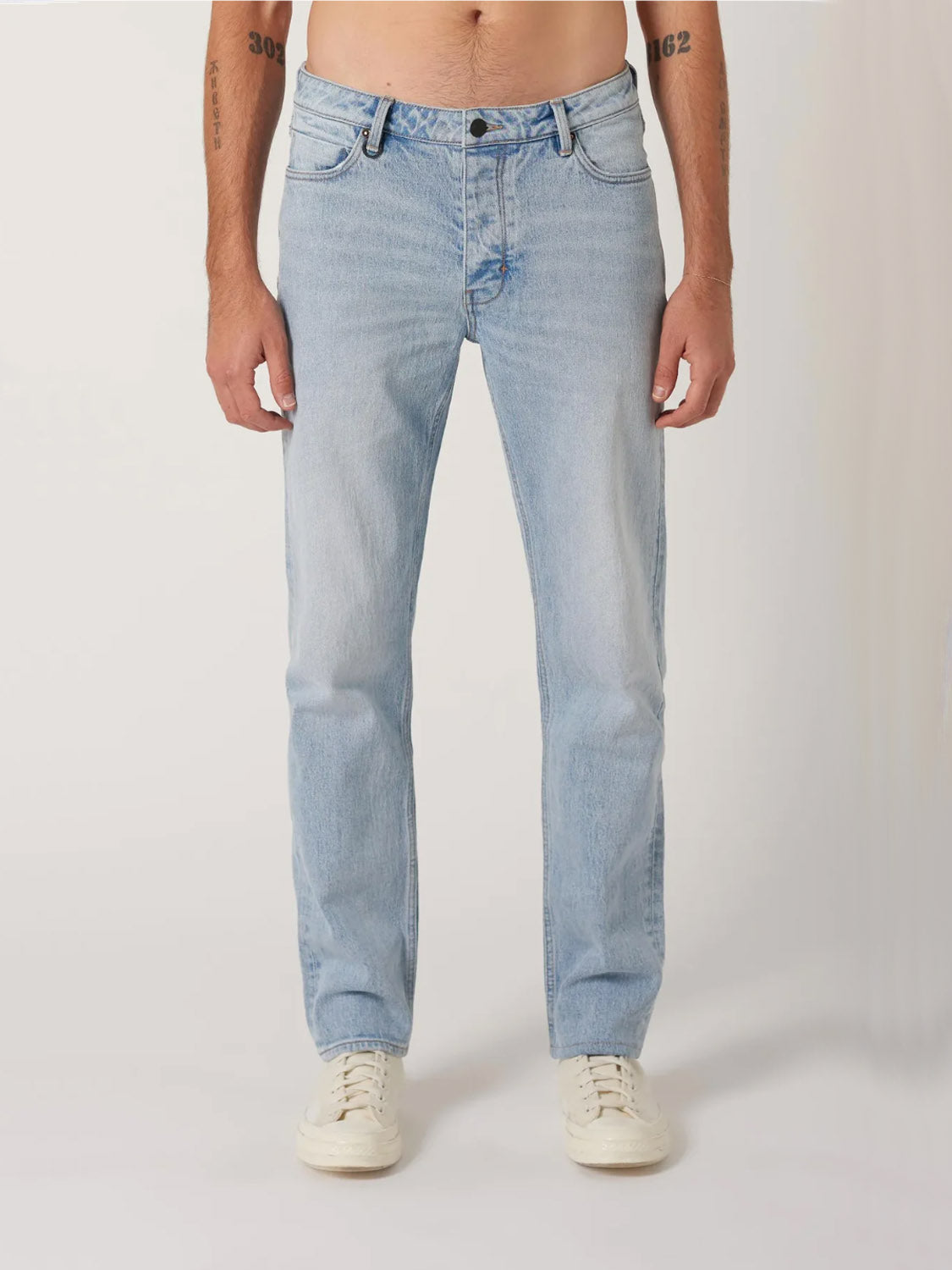 A person wears Neuw’s Ray Straight Baroque jeans in light blue with white sneakers, standing against a plain light background. The image is cropped at the torso with their arms relaxed at their sides.