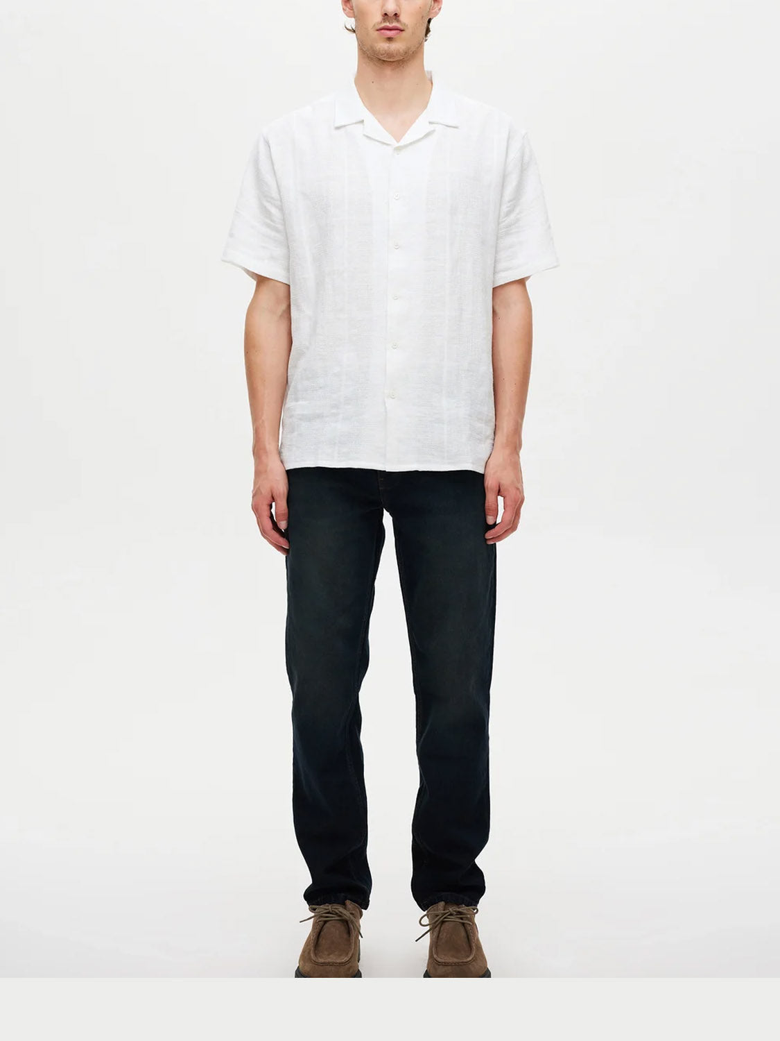 A person stands facing forward, wearing a white short-sleeve button-up shirt, Neuw Ray Straight Cosmo premium stretch denim jeans in dark blue, and brown suede shoes against a plain light background.