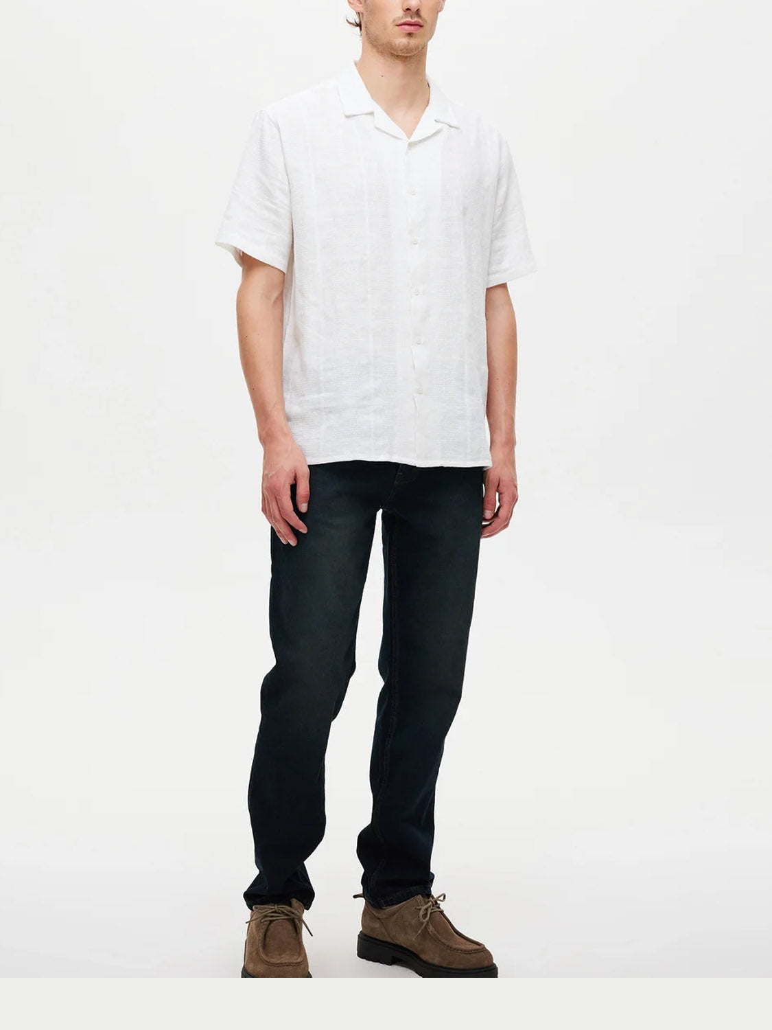 A man stands against a plain white background wearing a short-sleeve white button-up shirt, Neuw Ray Straight Cosmo premium stretch denim jeans in dark blue, and brown shoes.