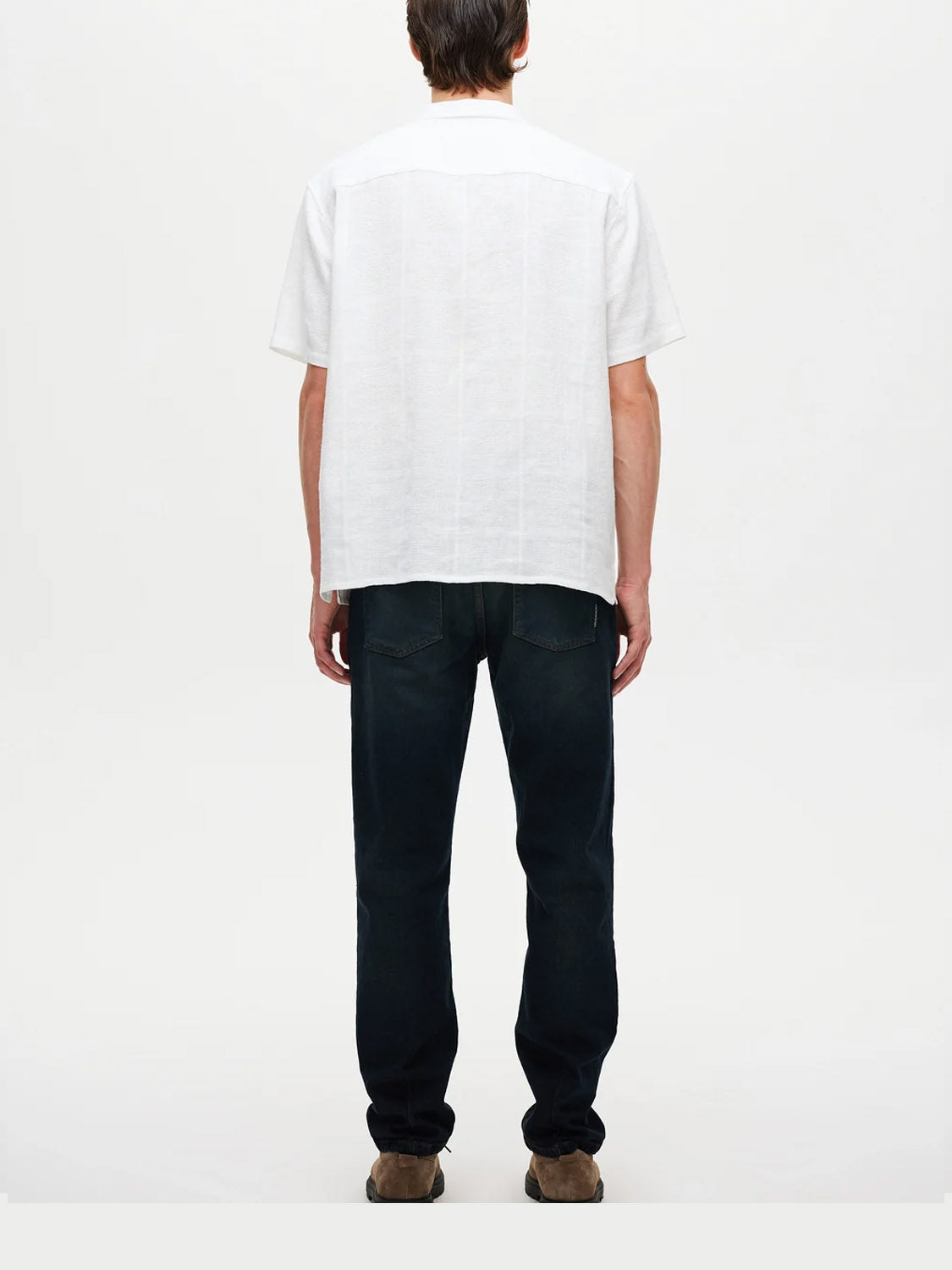 A person is facing away from the camera, wearing a white short-sleeve shirt and Neuw Ray Straight Cosmo dark blue jeans, paired with brown shoes against a plain light background.