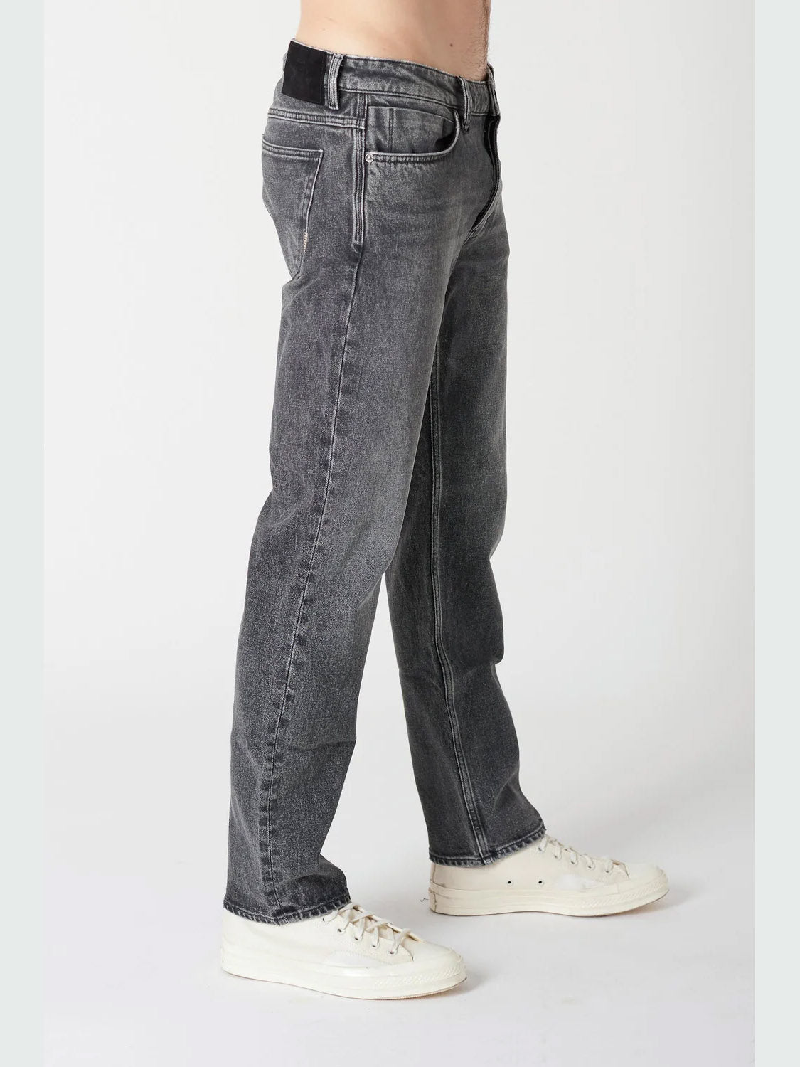 A person wearing Neuw Ray Straight Dead Moon jeans in a faded black wash with white sneakers stands against a plain light background, shown from the waist down in a side view.