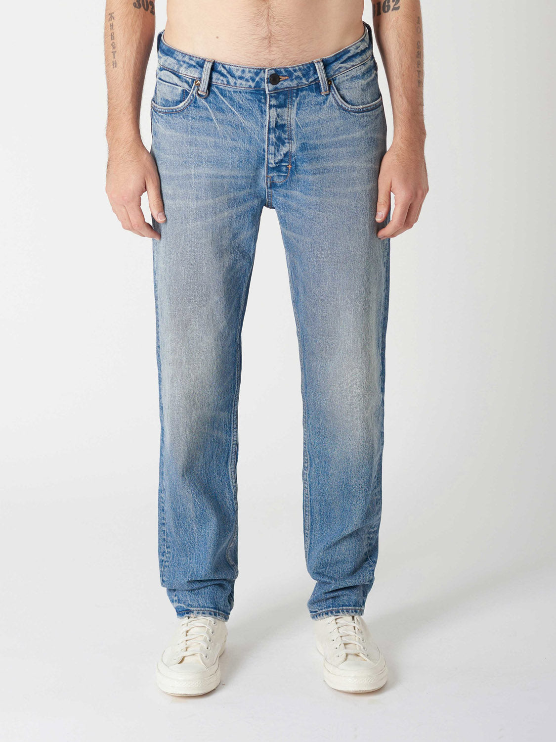 A person stands against a white background, wearing Neuw Ray Straight Decade jeans with a modern straight fit and white sneakers. Only the lower torso, legs, and arms are visible, hands relaxed at the sides.