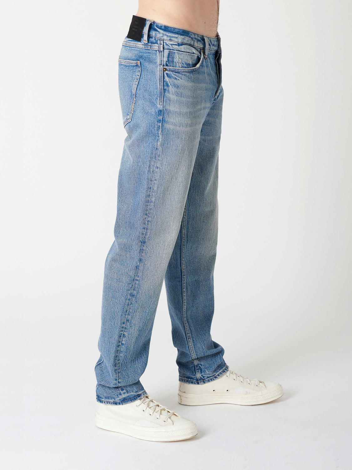 A person wears Neuw's Ray Straight Decade jeans with white sneakers, standing against a plain white background. Only their lower torso and legs are visible.