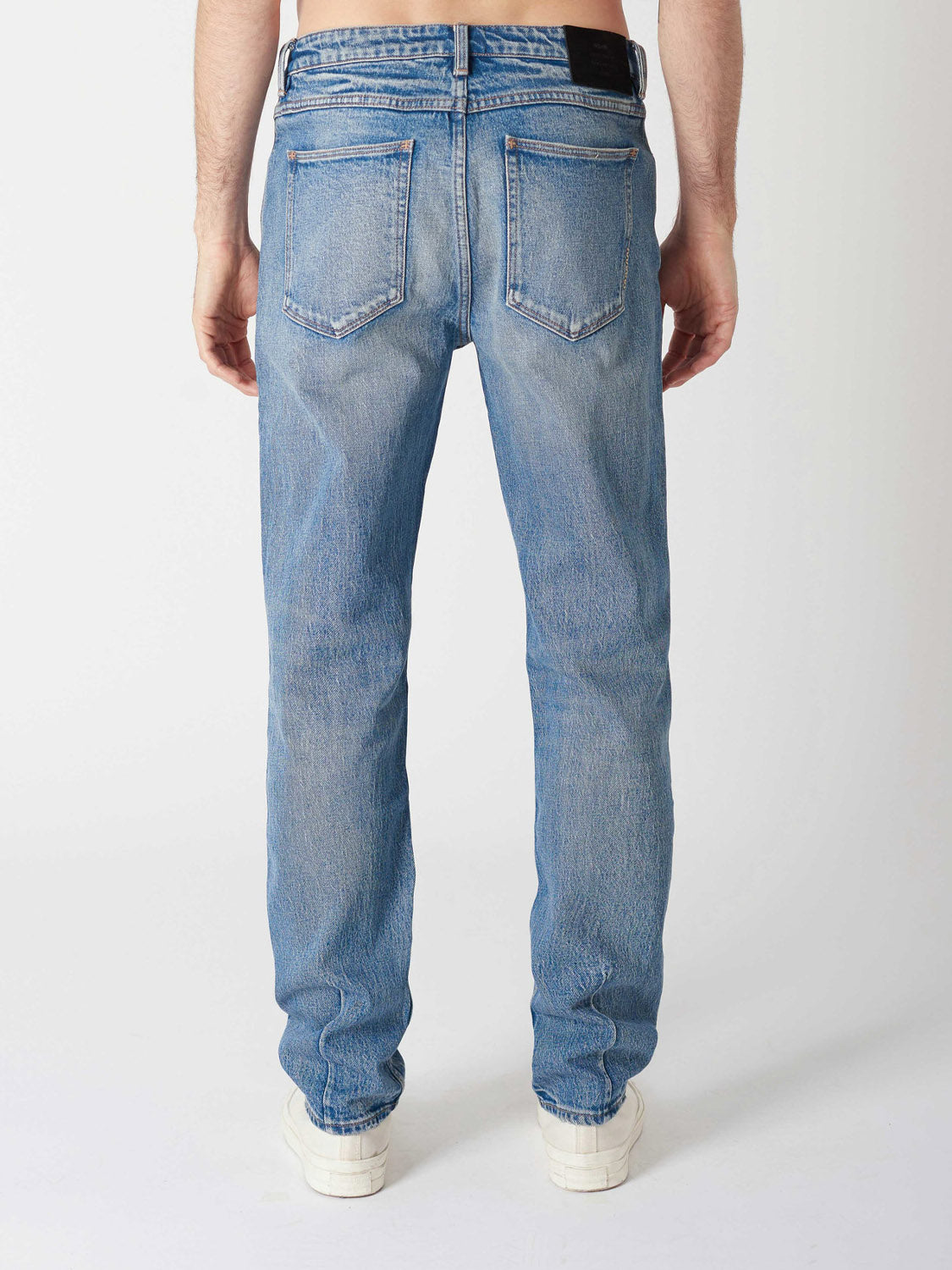A person is shown from the back wearing Neuw Ray Straight Decade light blue jeans with two back pockets and a leather patch on the waistband, paired with white sneakers, against a plain white background.
