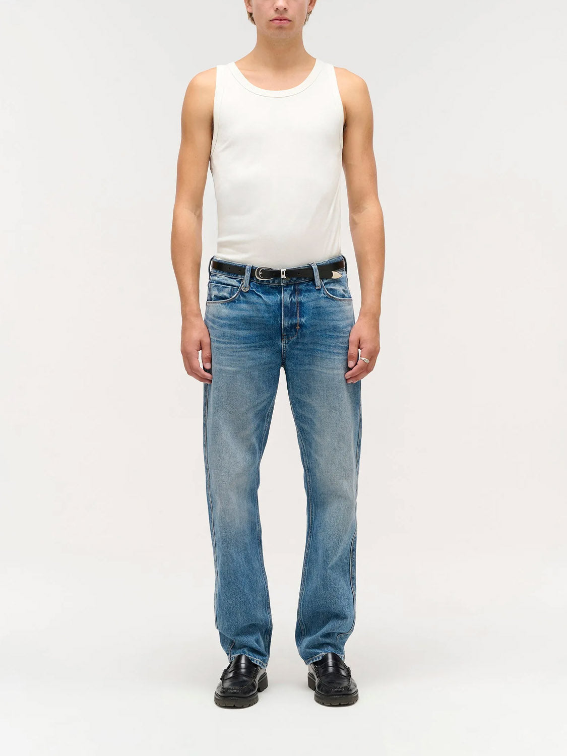 A person stands facing forward in a white sleeveless tank, Neuw Ray Straight Temper indigo jeans with a black belt, and black shoes against a plain light background.