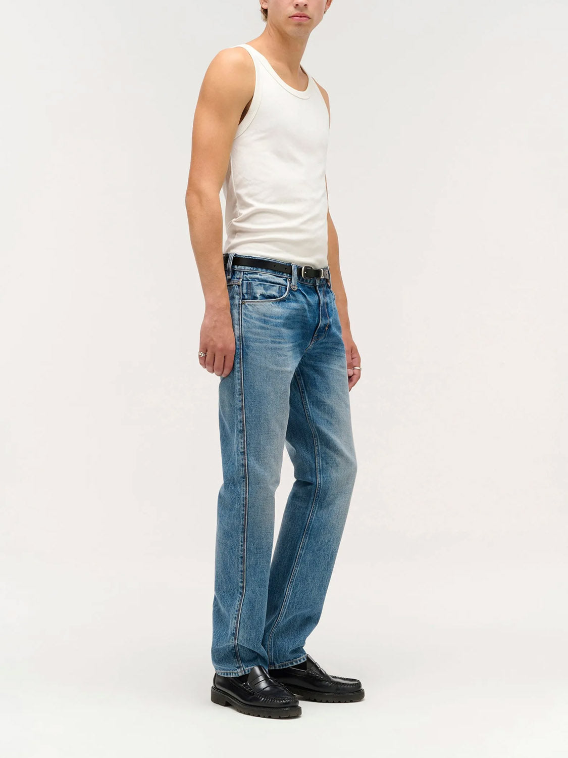 A person wears Neuw's Ray Straight Temper indigo denim jeans with a white tank top, black belt, and black shoes, standing against a plain light background. The face is partially out of frame.