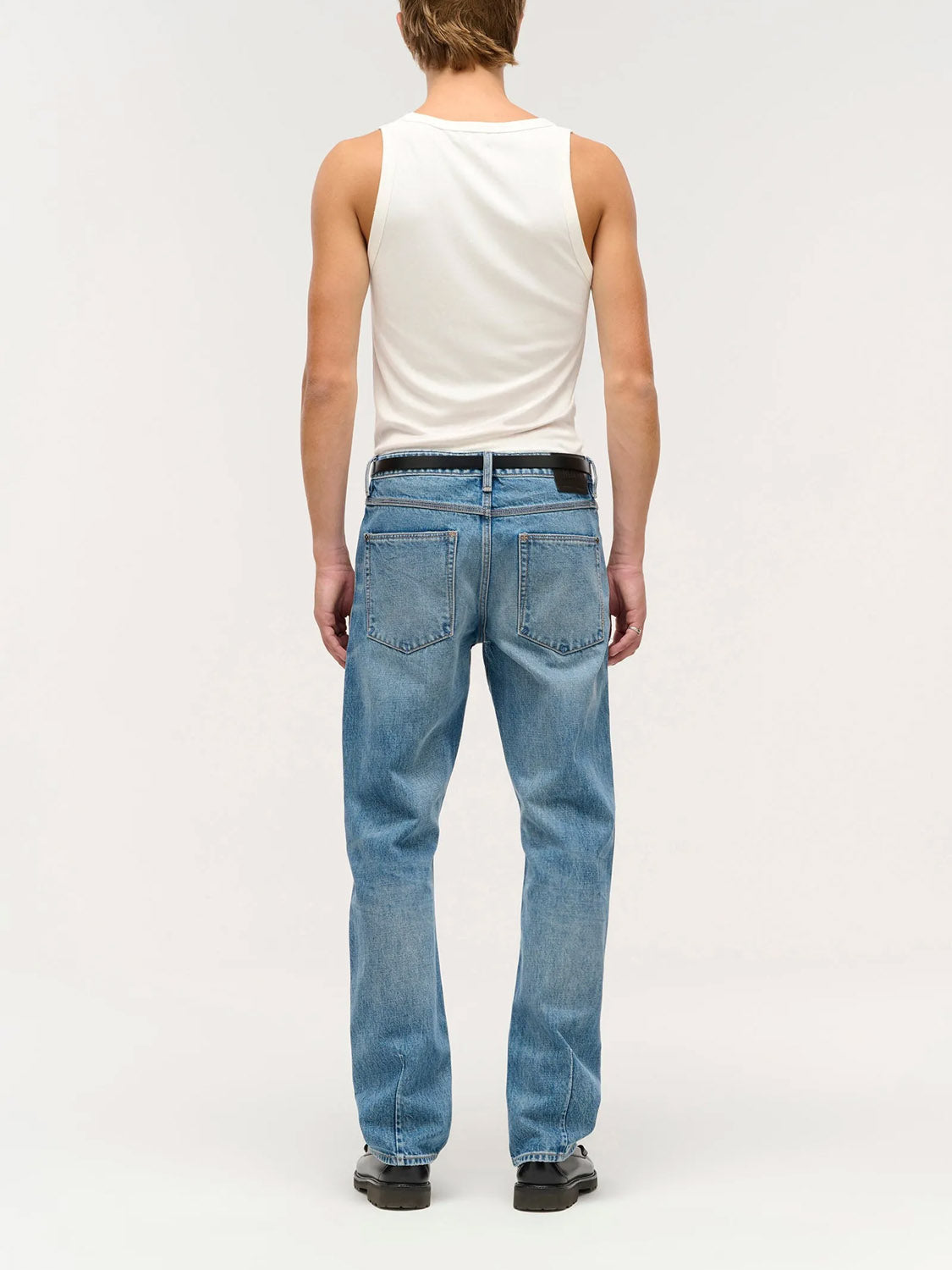 Someone with short hair, wearing a white sleeveless shirt, Neuw Ray Straight Temper light blue jeans, and black shoes stands facing away from the camera against a plain light background.