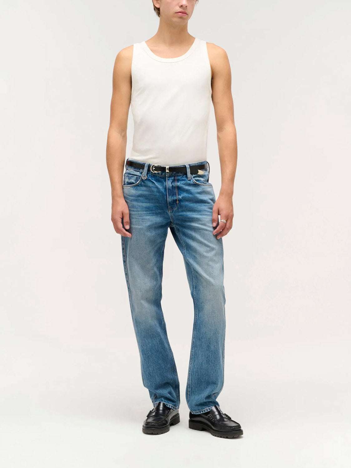 A person wears Neuw's Ray Straight Temper indigo denim jeans, a white tank top, black belt, and black shoes while standing against a plain light gray background.