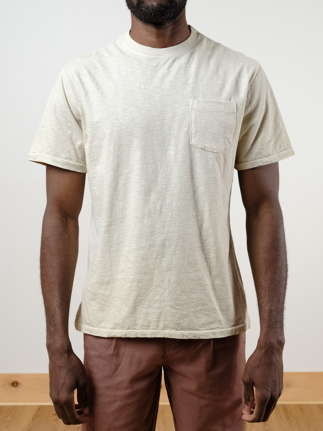 Someone models the ID Menswear Rebel Tee, a light beige slub cotton t-shirt with short sleeves and a chest pocket, paired with brown pants indoors against a plain light background. The shirt has a relaxed fit and the person&#39;s head is not shown.